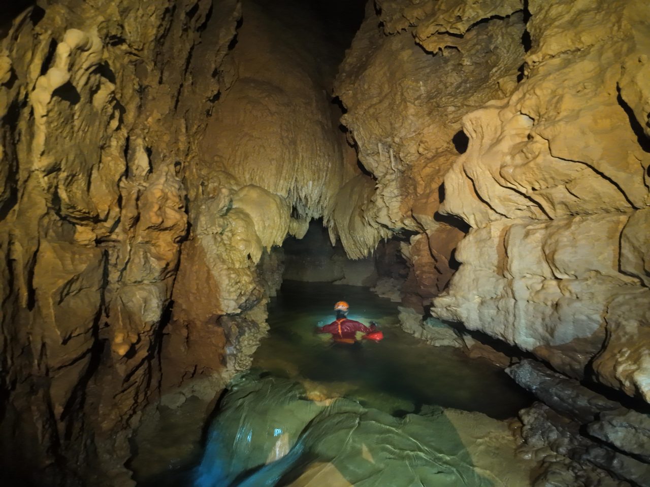Caver wading through a turquoise underground river beneath flowstone formations
